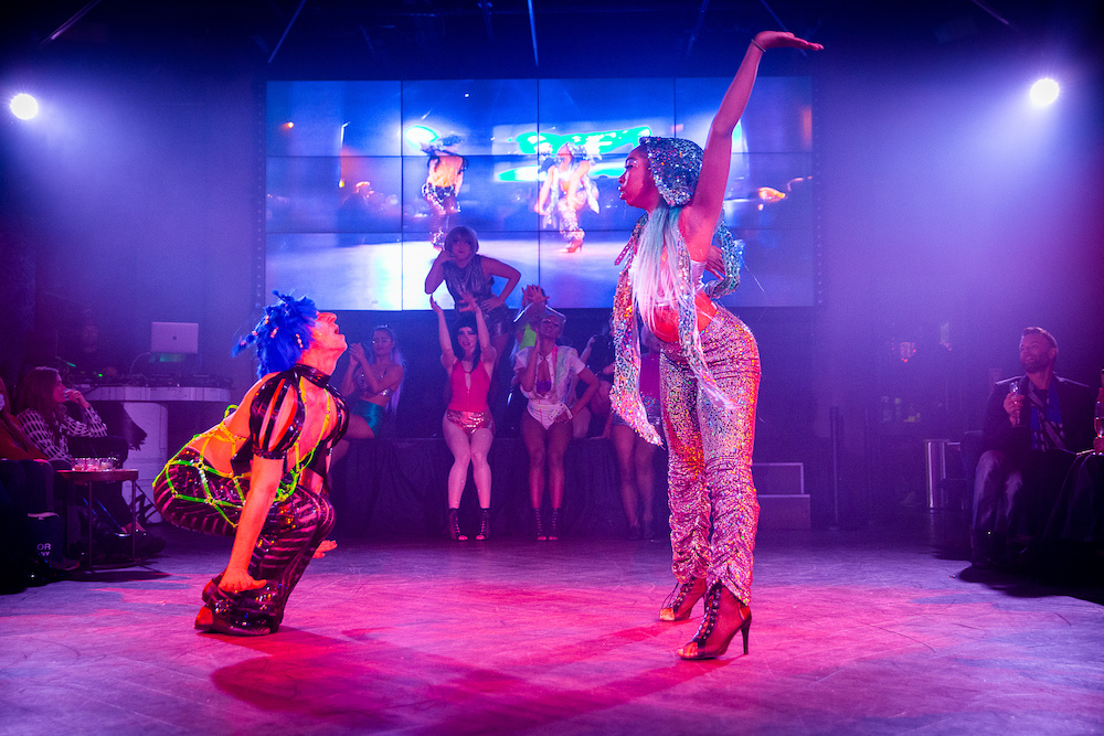 a club scene  people seated observe  a woman in a silver hooded, halter pantsuit dances with  a person in a blue headress, pink leopard pants and a neon-green net vest. 